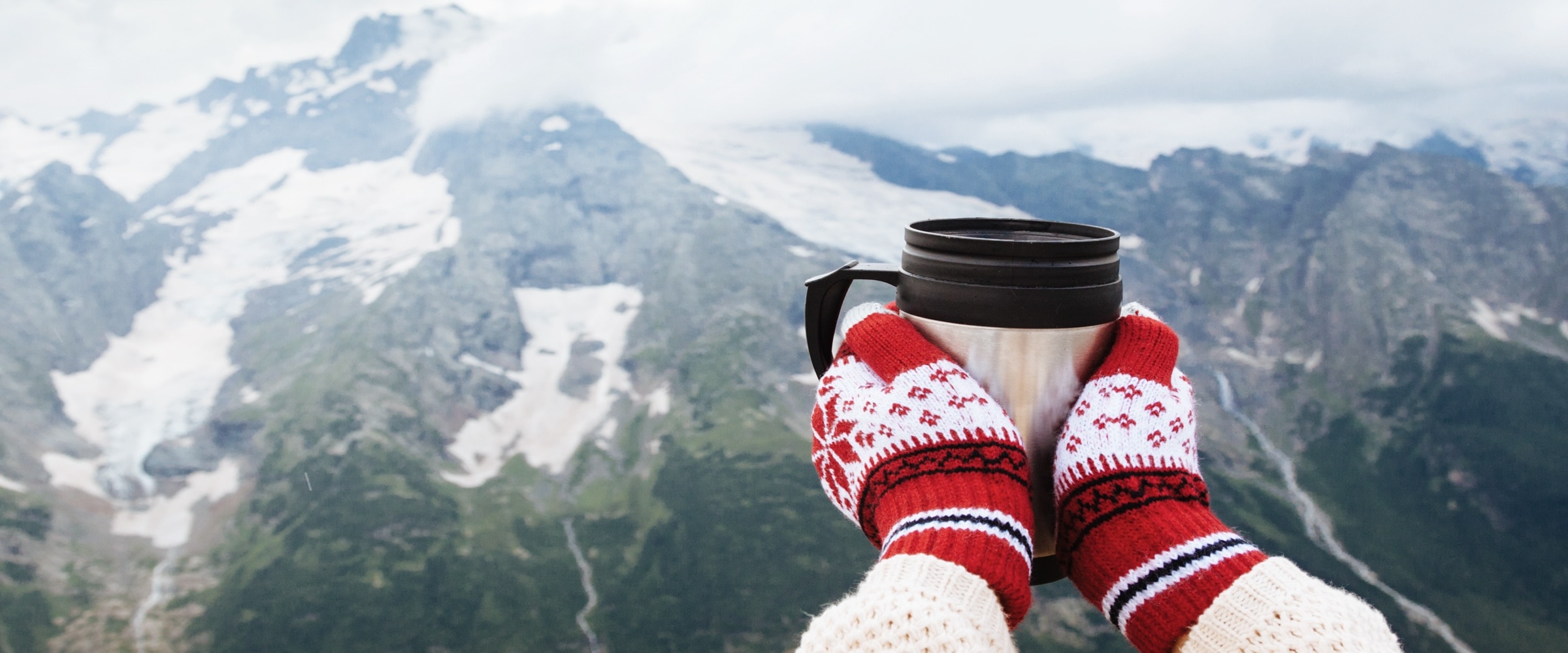 Photo of hands in mittens, holding a mug with a mountain in the background.