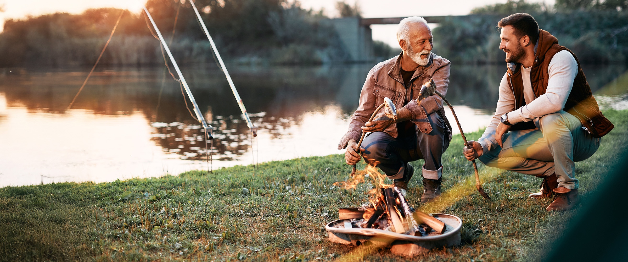 Photo of older and younger man squatting near campfire talking.