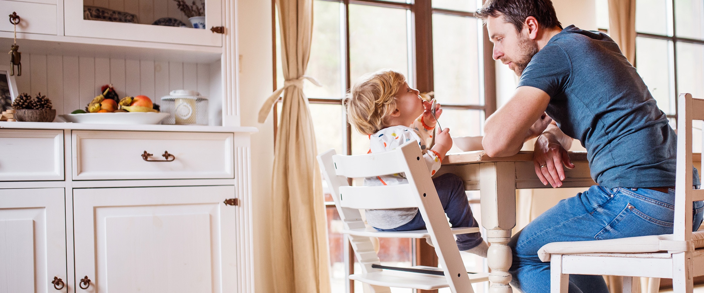 Photo of dad watching young toddler eat at the kitchen table.