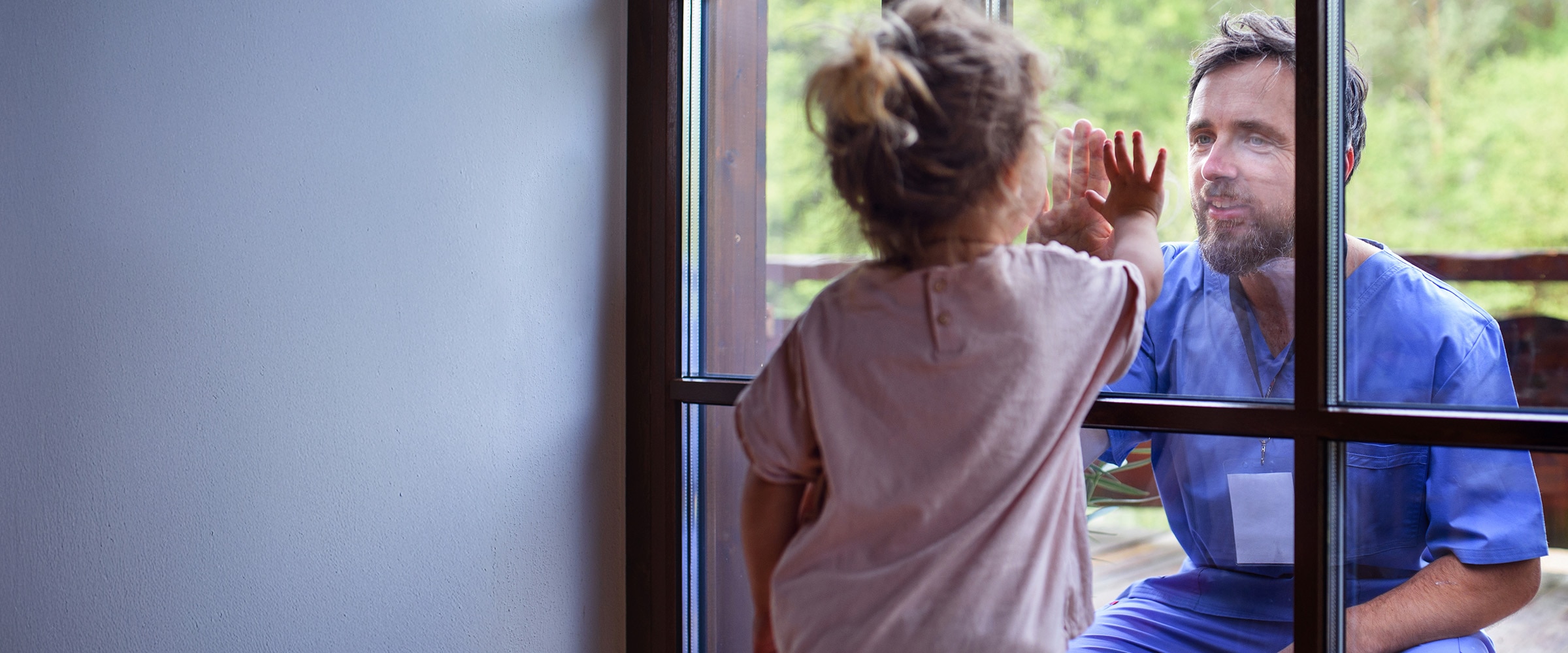 Photo of man in doctor scrubs, looking at young girl looking at him from the other side of the window.