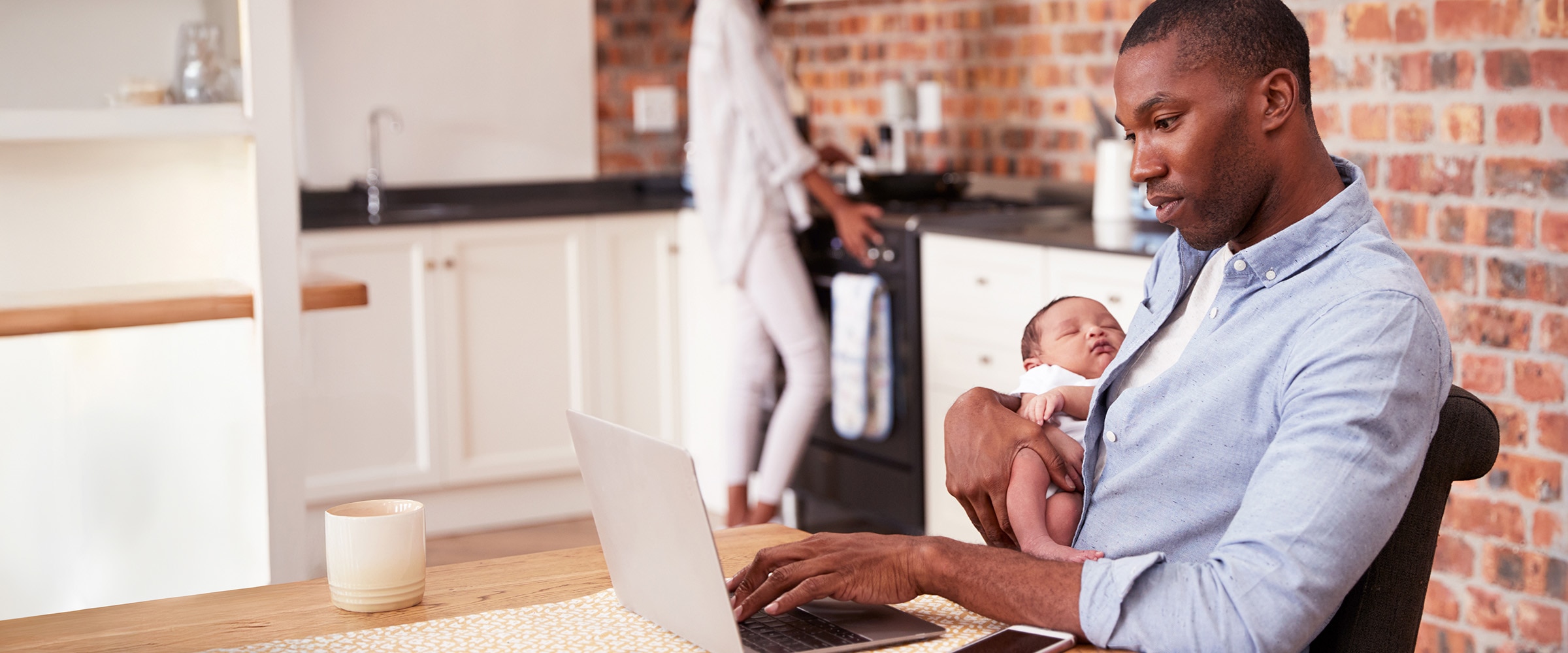 Photo of dad at table on a computer. He's holding a baby in one of his arms.