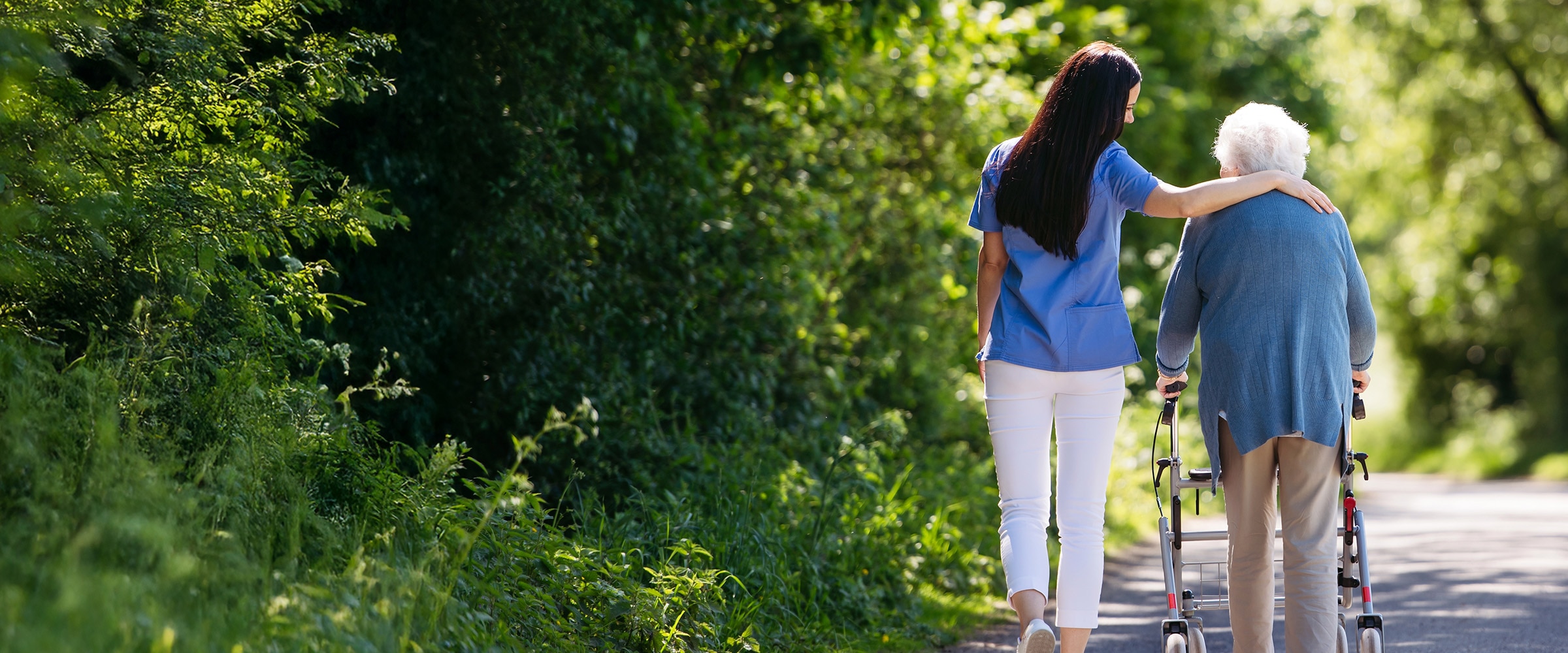 Photo of young woman walking with older woman down a tree-lined path.